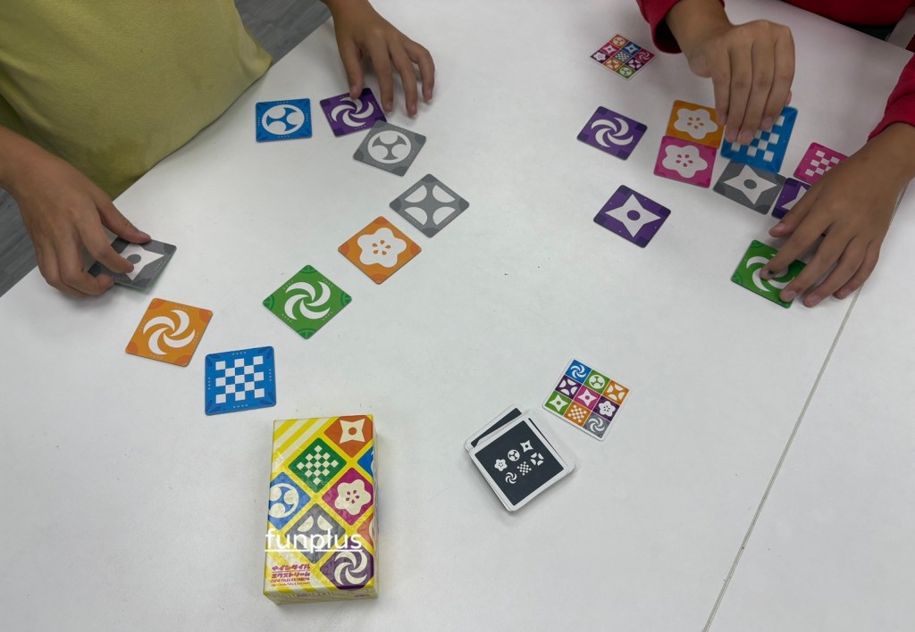兩隻手正在桌面上玩具卡牌遊戲,桌上有多種顏色的卡牌和一個遊戲盒,玩家正在擺放或選擇卡牌。